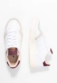 Vita sneakers med grå mockadetaljer och vinröda hältappar. Slät l äderöverdel, perforerad tå för ventilation och texturerad gummisula.