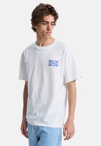 Νέος άνδρας που φοράει λευκό μπλουζάκι με ένα μικρό πολύχρωμο σχέδιο στο στήθος και ανοιχτόχρωμο μπλε τζιν, κοιτάζοντας προς τα αριστερά του.