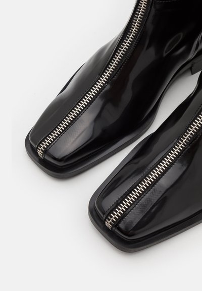 Svarta lackskinnankelstövlar har en fyrkantig tå och en silverfärgad dragkedjedetalj som löper ner fram. Designen är elegant och modern.