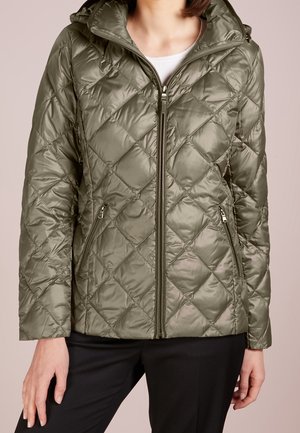 Kobieta w błyszczącej, oliwkowozielonej pikowanej kurtce z przednim zamkiem błyskawicznym, kieszeniami na zamek i kapturem, ubrana w czarne spodnie.