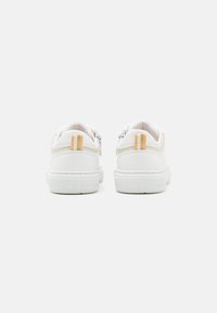 Zapatillas blancas con una parte superior de cuero liso, acentos de ante y rayas amarillas en el talón. Presentan una suela de goma texturizada con marca.