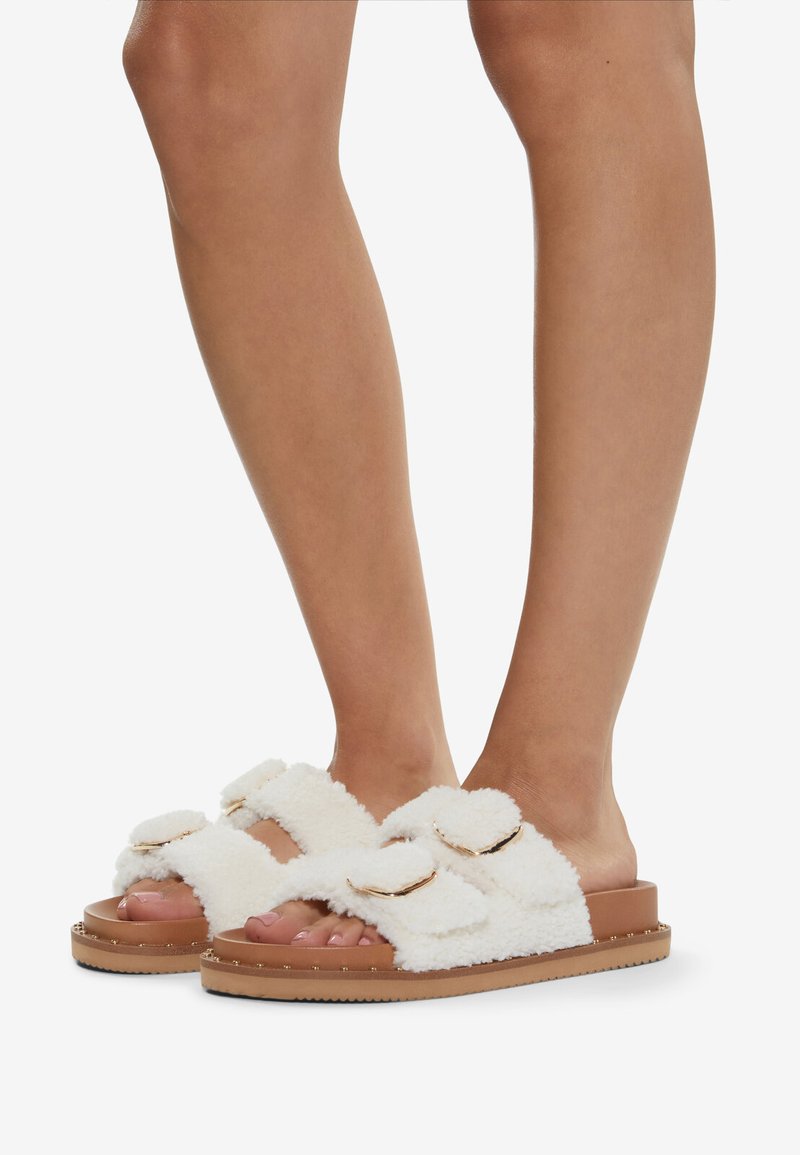 Witte fluffy slide sandalen met twee banden met gouden accenten, geplaatst op een tan-kleurige zool met een gestructureerd oppervlak en kleine studs langs de rand.