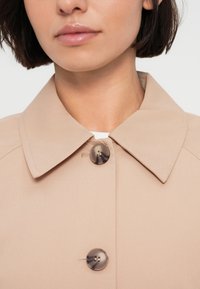 Παλτό με γιακά σε μπεζ χρώμα και λεία υφή, με μεγάλο κουμπί σε μοτίβο χελώνας και σχεδιασμό γιακά με δομή.