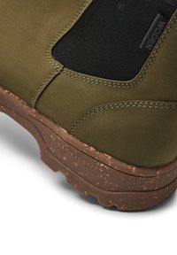 Botas verdes con un upper de tela suave, paneles laterales elásticos y un forro negro texturizado. Suela de goma marrón moteada marcada "NATURAL SOFT."