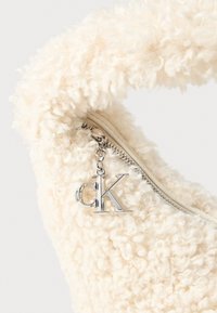 Crèmekleurige, pluizige tas met een gestructureerd oppervlak, ritsluiting en een zilveren bedeltje met "CK" eraan bevestigd.