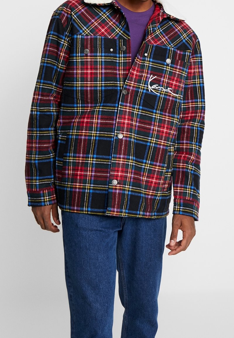 Rutig jacka i röd, svart och gul med en vit fårskinnskrage, tryckknappar och broderad logotyp på vänster bröstficka.