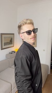 Jachetă bomber neagră cu guler ribbut, purtată peste o cămașă deschisă. Ochelari de soare negri. Păr de culoare deschisă. Canapea bej cu textură moale în fundal.
