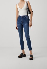 Džíny s vysokým pasem ve slim fit modré barvy, s minimálním poškozením. Kombinované s bílým topem s výstřihem do lodi a černými skluzovými sandály.