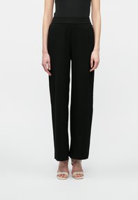 Kvinne iført svarte høy midje vide bukser og lyse beige åpne tå hæler, stående mot en plain hvit bakgrunn.