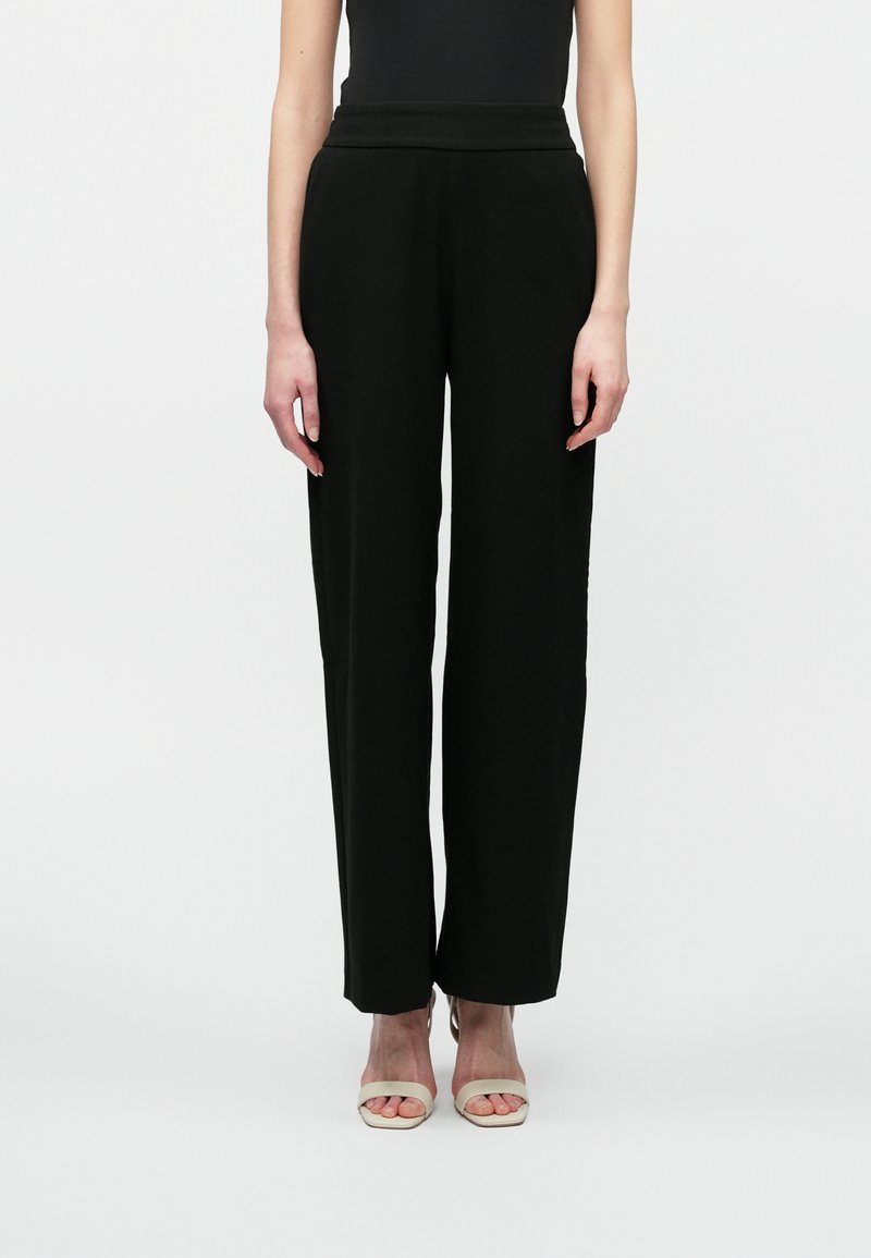 Kvinne iført svarte høy midje vide bukser og lyse beige åpne tå hæler, stående mot en plain hvit bakgrunn.