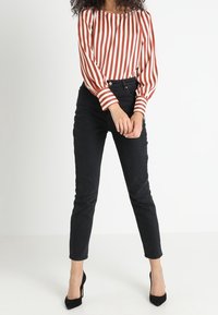 Rød og hvidstribet bluse med ballonsleeves, parret med sorte tætsiddende jeans. Sorte højhælede sko fuldender outfittet. Hvid baggrund.