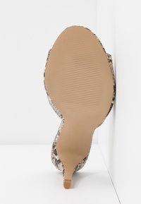 Semelle beige d'une chaussure à talon haut à motif peau de serpent, inclinée contre une surface blanche en angle.