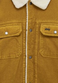 Giacca di velluto a coste in giallo senape con colletto in sherpa. Presenta due tasche sul petto, bottoni a pressione metallici e una trama verticale a coste.