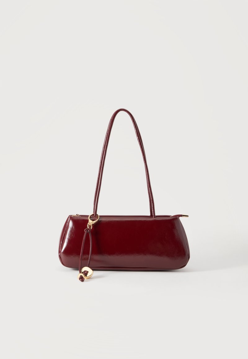 Sac à main en cuir rouge foncé brillant avec de longues anses et un charm circulaire doré suspendu à une anse.