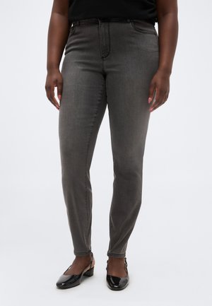 Persona che indossa jeans aderenti grigio scuro e scarpe slingback nere in vernice con tacco basso, in piedi davanti a uno sfondo semplice.