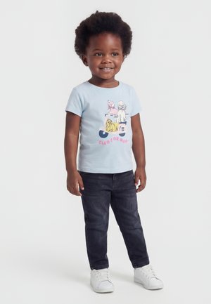 Niño pequeño sonriente con cabello rizado, usando una camiseta azul claro con estampado de perro y patinete, vaqueros oscuros y zapatillas blancas sobre un fondo blanco.