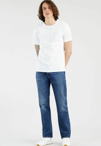 Witte katoenen t-shirt met reliëflogo, gecombineerd met blauwe denim jeans. Model draagt witte sneakers. Effen achtergrond, staande houding.