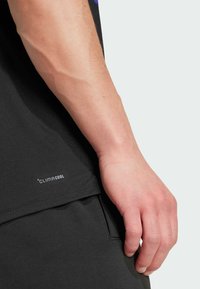 T-shirt noir à manches courtes avec un logo ClimaCool en bas. Gros plan d'une main reposant sur un pantalon sombre, mettant en valeur les textures et contours lisses.