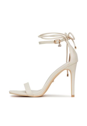 Sandalia blanca de tacón alto con lazos para atar en el tobillo, tira delgada sobre los dedos y detalle de hebilla en tono dorado sobre un fondo blanco.