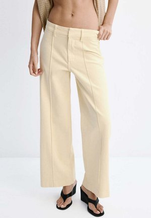 Beige wijde broek gedragen door een persoon met een blote buik, gecombineerd met zwarte sleehakken tegen een effen witte achtergrond.
