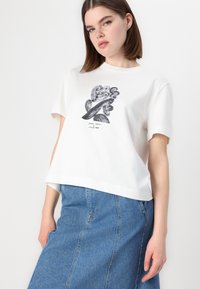 Ung kvinne i hvit T-skjorte med abstrakt svart design og tekst, kombinert med et høyt midjebånd blå denimskjørt mot en enkel bakgrunn.