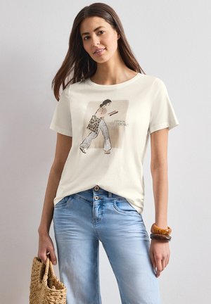 Kobieta w jasnobeżowym graficznym t-shircie i niebieskich dżinsach trzyma plecioną torbę z bransoletkami na nadgarstku na tle jednolitej ściany.
