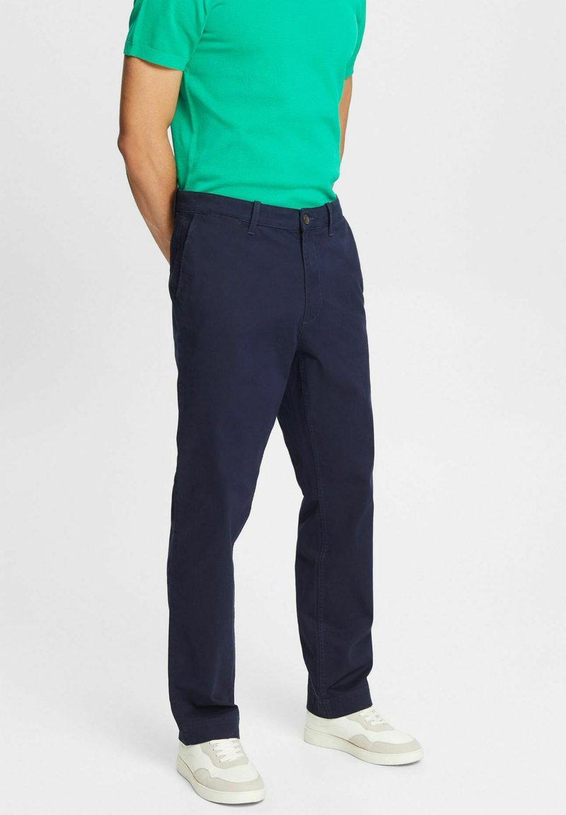 Vīrietis, kurš valkā tumši zilos bikses, baltas kedas un zaļu īsām piedurknēm krekls, stāv ar rokām aiz muguras uz vienkrāsaina fona.