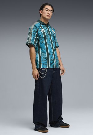 Uomo con occhiali, polo verde acqua con motivo a strisce verticali nere e bianche, jeans scuri a gamba larga con accessori a catena e scarpe nere.