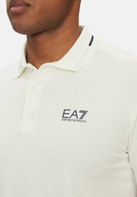 Polo en crema, confeccionado en tela texturada. Presenta un cuello con ribete en azul marino y un logo en azul marino en el lado izquierdo del pecho. Tres botones visibles.