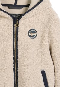 Roomkleurig fleecejack met marineblauwe biezen, ritssluiting en een ronde patch met een hond en de tekst "Marc O’Polo Friends" op de borst.