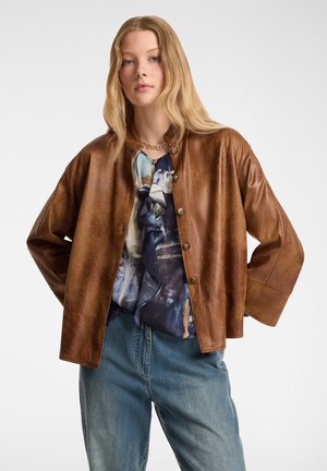 Bruine leren jas met een ronde halslijn en knoopsluiting, met ruime mouwen. Draagt over een blauw gebloemd blouse en jeans.