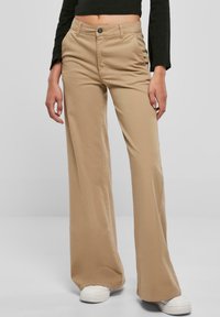 Beige wijde broek met een hoge taille, gemaakt van een gladde stof, met een knoopsluiting aan de voorkant en zijzakken. Witte sneakers zichtbaar.