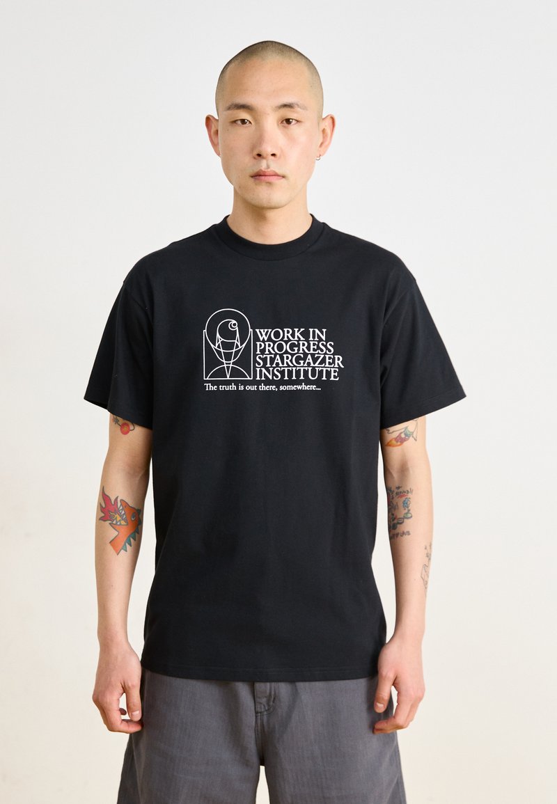 T-shirt preta de algodão com texto e gráficos brancos, mangas curtas, gola redonda, com tatuagens visíveis nos braços.