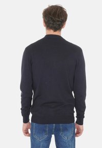 Uomo che indossa un maglione lungo nero semplice e jeans blu, visto da dietro su uno sfondo bianco.