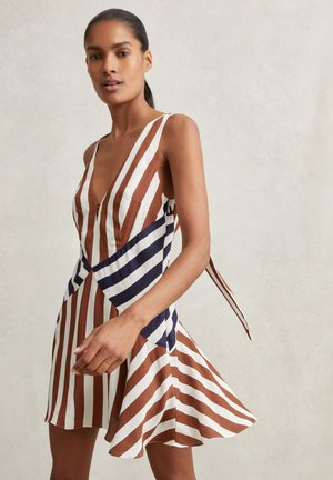 Femme portant une robe sans manches rayée marron, blanche et bleu marine avec un col en V et une jupe évasée sur un fond clair uni.