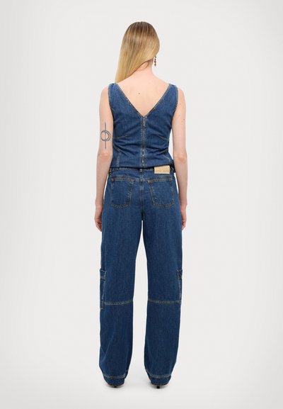 Combinaison en denim avec un dos en V, un design à jambes larges, deux poches et des coutures contrastées. Le tissu est bleu avec une texture classique.