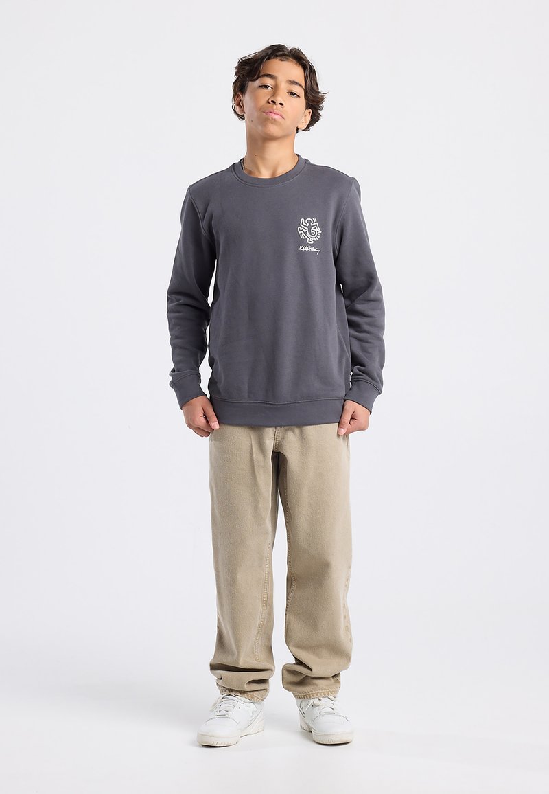 Sudadera gris con cuello redondo y logo frontal, combinada con pantalones beige y zapatillas blancas, sobre un fondo neutro.