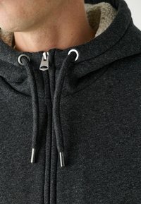 Dunkelgraue Zip-Hoodie mit einem weichen, beigen Fleece-Futter. Verfügt über einen metallischen Reißverschluss und Zugstopper sowie strukturierter Stoff für zusätzliche Wärme.