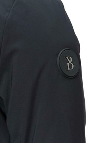 Czarna kurtka o gładkiej fakturze, z okrągłą czarną łatą na rękawie, na której widnieje stylizowana litera "B". Prosty, nowoczesny design.