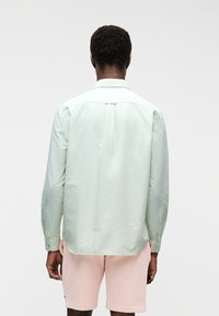 Personne aux cheveux courts et bouclés, vue de dos, portant une chemise à manches longues vert clair et un short rose pâle sur un fond uni.