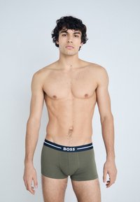 Olívazöld boxeralsó fekete-fehér márkás derékpánttal. Az anyaga simának és testhezállónak tűnik, rövid lábakkal és minimális varrásokkal.