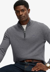 Maglione grigio con zip, colletto a coste, texture in maglia e piccolo logo sul petto, indossato sopra una camicia bianca e abbinato a jeans blu.