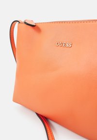 Sac en cuir orange à porter en bandoulière avec une fermeture éclair, arborant un logo "GUESS" en or et une sangle noire contrastante. Texture lisse avec un design simple.