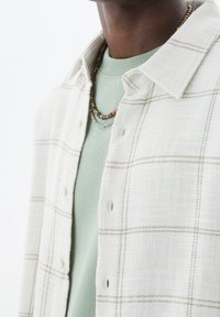 Camisa de cuadros crema con líneas marrones claras, superpuesta a un suéter verde apagado. Presenta botones y tejido texturizado. Accesorios con collares de cuentas.