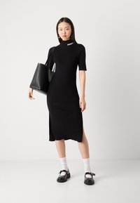 Abito nero in maglia a coste con collo alto e spacco laterale, abbinato a borsa e scarpe nere, accentuato da calzini bianchi. Presenta il logo DKNY.