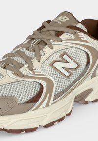 Μπεζ και καφέ παπούτσι τρεξίματος New Balance με λεπτομέρειες από δίχτυ και δέρμα, με χοντρή σόλα και μπεζ κορδόνια δεμένα σε φιόγκο.