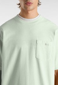 Hellgrünes Baumwoll-T-Shirt mit rundem Halsausschnitt, kurzen Ärmeln und einer Brusttasche mit einem kleinen Markenetikett. Glatte Textur.