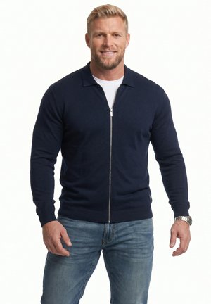 Mann mit kurzem blondem Haar und Bart, der einen marineblauen Pullover mit Reißverschluss, ein weißes Hemd, blaue Jeans und eine Armbanduhr trägt, lächelnd und stehend.