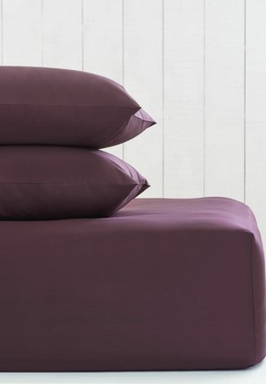 Due cuscini viola scuro impilati su un lenzuolo aderente abbinato contro una parete di pannelli in legno bianco.