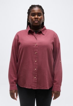 Žena s vrkočmi vo vlasoch, ktorá má na sebe zlaté kruhové náušnice, zapínanú vínovo červenú košeľu a čierne nohavice, stojaca na jednoduchom svetlom pozadí.